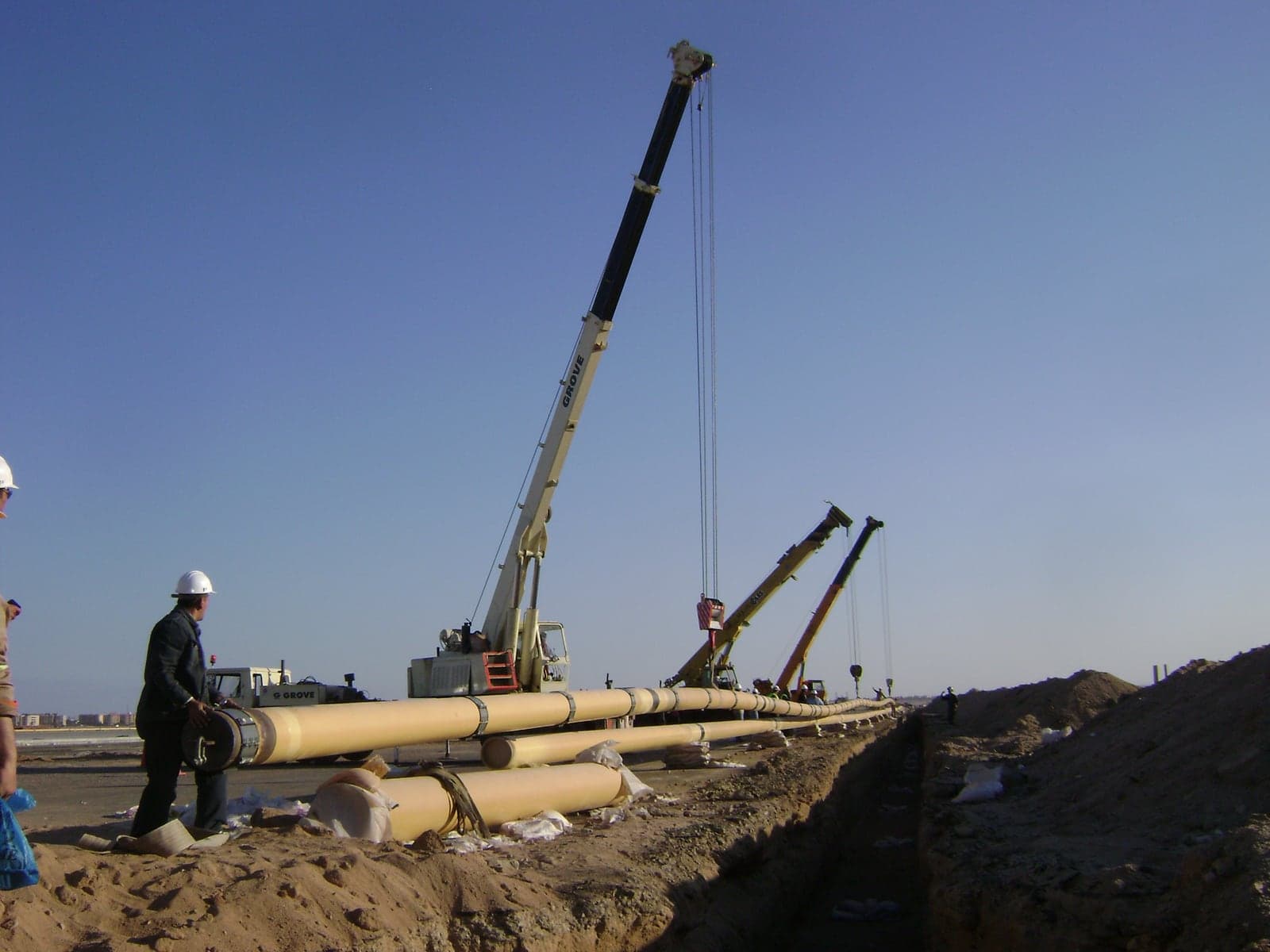 Cranes stringing large-diameter hydrant mains beside an excavated trench