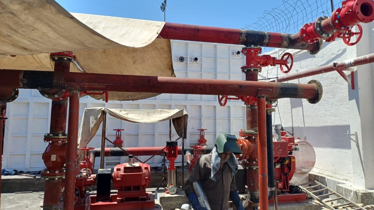 Overhead red fire mains and gate valves above pump sets under a tarpaulin, a fitter working between them