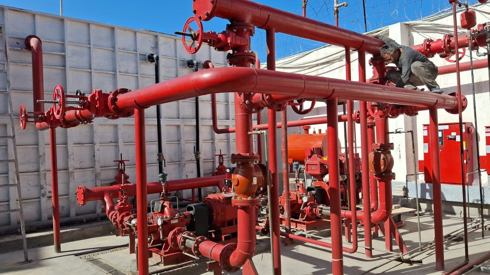 Red fire-fighting pipework with gate valves and flanged headers over a pump set in a walled yard, an orange pressure vessel and red controller cabinets behind